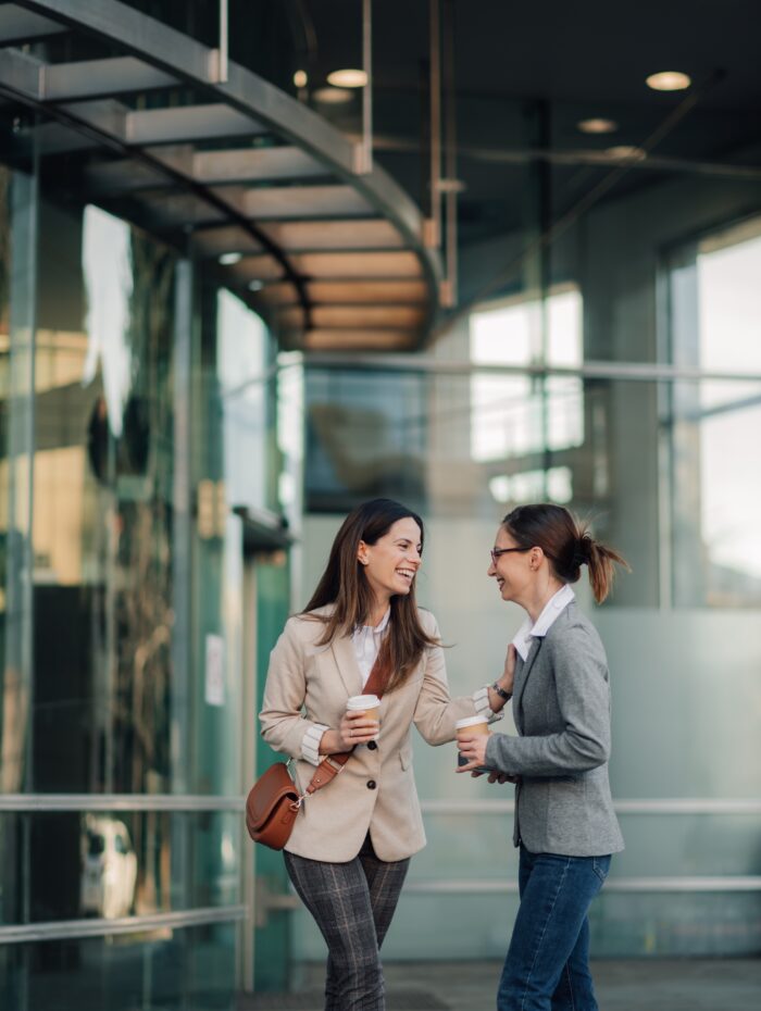 Two businesswomen enjoying a coffee break outside the office, engaging in lively conversation and sharing laughter while fostering a positive and supportive relationship in a vibrant urban setting